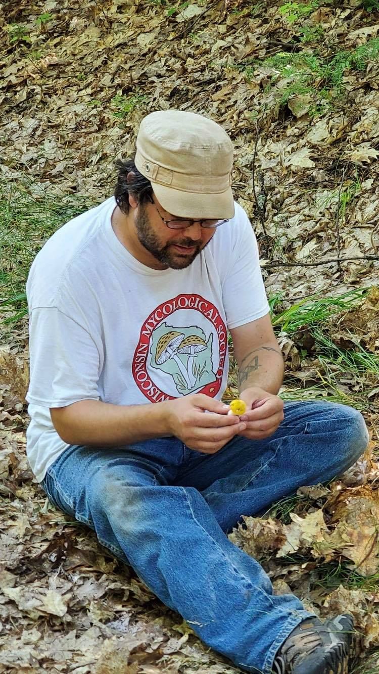 Tavis Lynch wearing a Wisconsin Mycological Society T-Shirt and a baseball cap, looking down at a mushroom, looks like a Chanterelle.