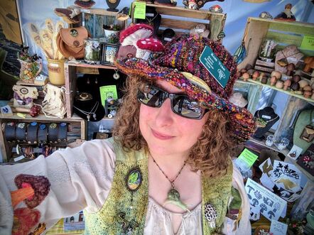 Rose Tursi wearing a funky hat she made with two fabric Amanita muscaria shrooms on it. She also has on sunglasses and is in a studio area with multiple art projects in the background.